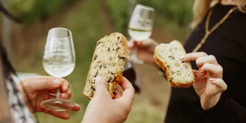 Zwei Personen halten Gläser mit Weißwein und Stücke von frisch gebackenem Brot. Im Hintergrund sind grüne Weinreben zu sehen.