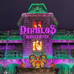 Ein gruseliges, beleuchtetes Gebäude mit dem nächtlichen Schriftzug "DIABLOS RESIDENZ". Farbige Lichter in Violett und Grün schaffen eine geheimnisvolle Atmosphäre.