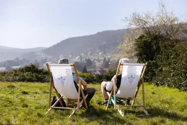 Zwei Personen sitzen in Liegestühlen auf einer Wiese und genießen den Ausblick auf eine malerische Landschaft. Die Sonne scheint und der Himmel ist klar.