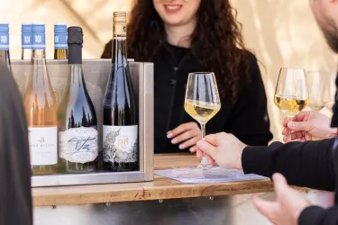 Eine Weinbar mit verschiedenen Flaschen und Gläsern. Eine Person reicht ein Glas Wein und eine weitere Person steht am Tisch.