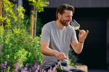 Ein Mann genießt ein Glas Wein in einer grünen Umgebung. Er sitzt entspannt und freut sich über den Moment.