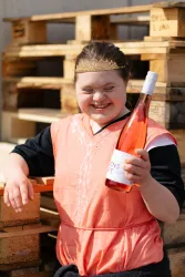 Eine fröhliche Person hält eine Flasche Roséwein in der Hand. Im Hintergrund sind Holzpaletten zu sehen.