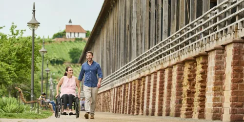 Ein Paar geht an einer langen Mauer entlang, während die Frau im Rollstuhl sitzt. Die Umgebung ist grün und ein Haus ist im Hintergrund sichtbar.