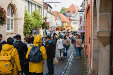 Eine belebte Straße mit vielen Menschen und bunten Gebäuden. Blumen hängen von den Fenstern, und die Atmosphäre ist lebhaft.