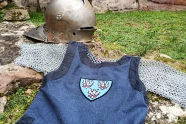 Ein mittelalterlicher Helm liegt neben einem blauen Wappenrock mit einem Emblem auf dem Boden.