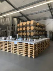 Eine moderne Weinlagerung mit Holzfässern und einem langen Tisch aus Paletten. Darauf stehen verschiedene Weinflaschen.