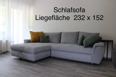 Ein modernes Schlafsofa in Grau steht in einem hellen Raum. Es hat eine Liegefläche von 232 x 152 cm und bietet gemütliche Kissen.