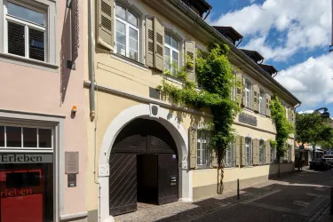 Ein historisches Gebäude mit grüner Bepflanzung und Fensterläden. Die Straßenansicht zeigt eine charmante Fassade und einen Eingang mit einem großen Tor.