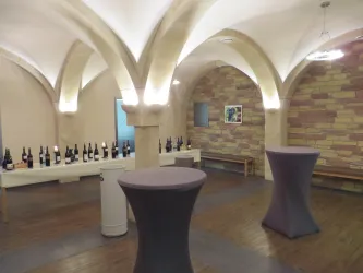 Ein Weinkeller mit eleganten Gipsbögen und Holzfußboden. Auf dem Tisch stehen viele Flaschen Wein, daneben gibt es Stehtische.