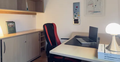 Ein moderner Büroarbeitsplatz mit einem Schreibtisch und einem Laptop. Der Raum ist hell und enthält einige Bücher sowie dekorative Elemente an der Wand.
