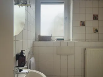 Ein Badezimmer mit weißen Fliesen und einem großen Fenster. Auf der Wand hängen einige kleine Bilder und es gibt eine Handtuchhalterung sowie einen Heizkörper.