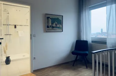 Ein helles Zimmer mit einem Blick auf eine Stadt und einem bequemen Sessel. Im Hintergrund ist ein Badezimmer mit einer modernen Dusche sichtbar.