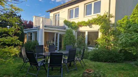 Ein modernes Haus mit großen Fenstern und einem gepflegten Garten. Im Vordergrund steht ein Tisch mit Stühlen, umgeben von grünen Pflanzen.