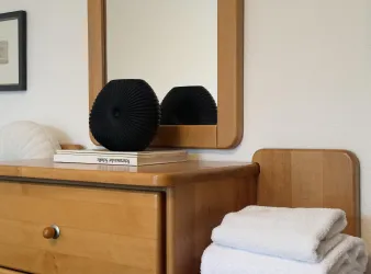 Ein modern eingerichteter Nachttisch mit einem Holzschrank, einem Spiegel und einer schwarzen Vase. Neben der Vase liegen weiße Handtücher und ein Buch.