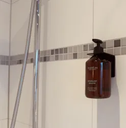 Ein braunes Duschgel in einem modernen Spender hängt an einer weißen Fliese in der Dusche. Die Fliese hat eine dekorative Bordüre aus grauen Mosaiksteinen.