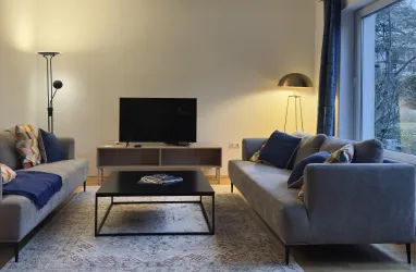 Ein modernes Wohnzimmer mit zwei Sofas, einem Tisch und einem Fernseher. Das Licht ist warm und der Raum hat große Fenster, die nach draußen blicken.
