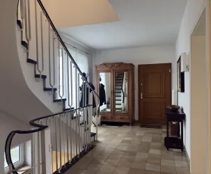 Ein heller Flur mit einer geschwungenen Treppe und einem eleganten Spiegel. An der Wand hängen Jacken, und eine Holzkommode steht neben der Eingangstür.