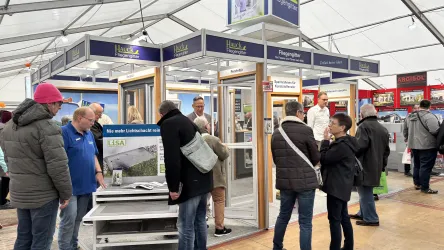 Eine Gruppe von Menschen diskutiert an einem Stand über Treppen und Holzarbeiten. Im Hintergrund sind verschiedene Holztreppen und Informationsplakate zu sehen.