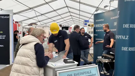 Eine Messe mit mehreren Personen, die an einem Stand stehen und sprechen. Im Vordergrund steht eine Frau, die an einem Laptop arbeitet.
