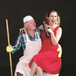 Ein Mann in Kochkleidung sitzt mit einem fröhlichen Lächeln auf einer Frau in einem roten Kleid. Sie halten verschiedene Utensilien und lachen zusammen.