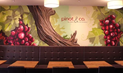 Ein gemütliches Restaurant mit einer Wandmalerei von Trauben und Weinblättern. Die Holztische und der moderne Stil schaffen eine einladende Atmosphäre.