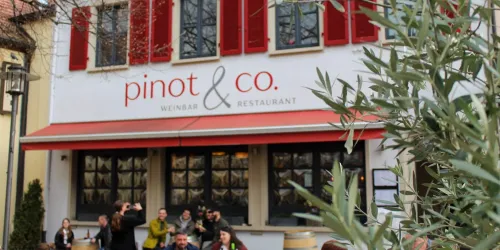 Ein Restaurant mit dem Namen „pinot & co.“ ist zu sehen. Vor dem Gebäude stehen mehrere Menschen, und die Fenster sind mit roten Fensterläden geschmückt.