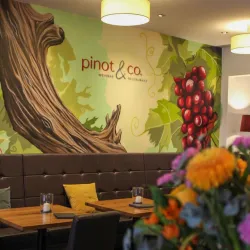 Ein stilvolles Restaurant mit einer Wandmalerei von Weinreben und Trauben. Es gibt gemütliche Sitzbänke und Tische aus Holz.