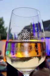 Ein Glas mit Weißwein ist im Vordergrund zu sehen. Im Hintergrund leuchten bunte Lichter und Menschen sind zu erkennen.