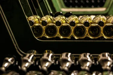 Eine Reihe von Flaschen mit goldenen und schwarzen Kappen ist ordentlich gestapelt. Im Hintergrund sind weitere Flaschen sichtbar, die in einem dunklen Raum aufbewahrt werden.