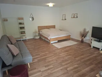 Ein modernes, helles Schlafzimmer mit einem Doppelbett und einem gemütlichen Sofa. Der Raum hat einen warmen Holzfußboden und dezente Dekoration.