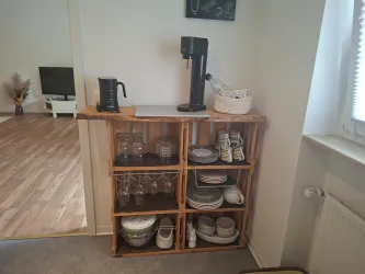 Ein Holzregal mit Geschirr und Gläsern in einer hellen Wohnung. Oben steht eine Kaffeemaschine neben einer Schüssel mit Zubehör.