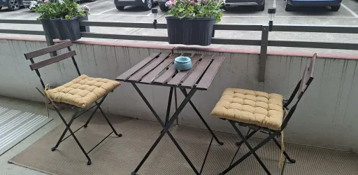 Ein kleiner Balkon mit einem Tisch und zwei Stühlen. Auf dem Tisch steht eine dekorative Schale und Blumenkästen sind im Hintergrund zu sehen.