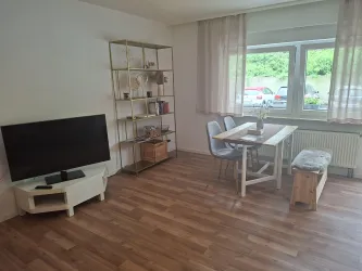 Ein gemütliches Wohnzimmer mit einem Fernseher, einem Esstisch und Stühlen. Die Fenster bieten einen Blick ins Freie und lassen viel Licht herein.