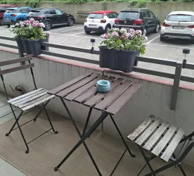 Ein kleiner Balkon mit zwei Holztischen und Stühlen. Auf der Brüstung stehen blühende Pflanzen in Kasten.