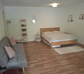Ein helles Schlafzimmer mit einem großen Bett und einem gemütlichen Schlafsofa. Der Raum hat Holzfußboden und einfache, moderne Möbel.