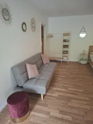 Ein modernes Wohnzimmer mit einem grauen Sofa und zwei Kissen. Es gibt einen runden Hocker in Rot und goldenen Akzenten sowie dekorative Spiegel an der Wand.