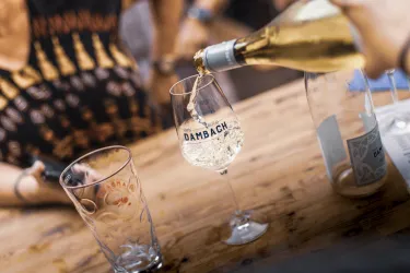 Ein Glas wird mit Weißwein gefüllt, während ein weiteres leeres Glas daneben steht. Im Hintergrund sind unscharfe Hände und ein Tisch zu sehen.