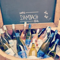 Eine Holztonne voller Weinflaschen mit Eis und einer Schiefertafel im Hintergrund. Die Tafel trägt die Aufschrift "Dambach" und "Zum Wohl!".