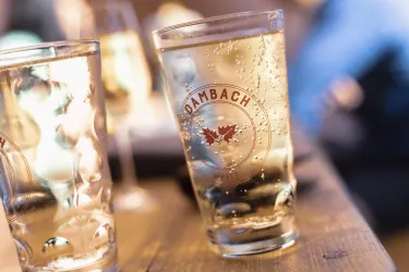 Ein Glas mit sprudelndem Getränk steht auf einem Holztisch. Im Hintergrund sind verschwommene Personen zu sehen.