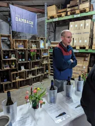 Ein Mann steht an einem Tisch mit Weinen in einer Lagerumgebung. Im Hintergrund sind Kisten und eine Banner-Werbung sichtbar.