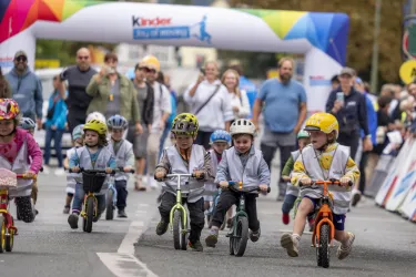 Eine Gruppe von Kindern fährt auf kleinen Fahrrädern. Im Hintergrund sind viele Zuschauer und ein buntes Zielbanner zu sehen.