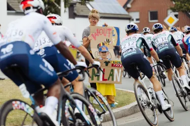 Eine Gruppe von Radfahrern fährt vorbei, während eine Frau mit einem Schild steht. Neben ihr steht ein Kind, das lächelt und die Zuschauer anfeuert.