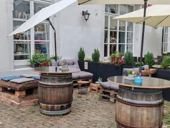 Ein einladender Außenbereich mit runden Tischen aus Holzfässern und bequemen Sitzgelegenheiten. Bunte Pflanzen und Sonnenschirme schaffen eine gemütliche Atmosphäre.