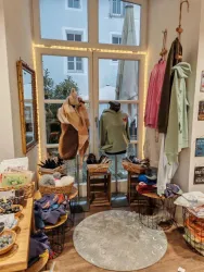 Ein gemütlicher Raum mit Kleidung und Accessoires auf Holzregalen. Durch das Fenster scheint Tageslicht und schafft eine einladende Atmosphäre.