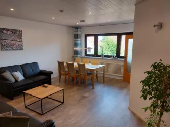 Ein gemütlicher Wohnraum mit einem schwarzen Sofa, einem Holztisch und Stühlen. Große Fenster sorgen für viel Licht und eine einladende Atmosphäre.