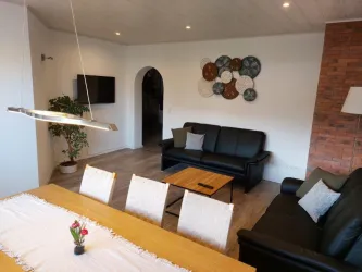 Ein modernes Wohnzimmer mit einem schwarzen Sofa und einem Esstisch. An der Wand hängen dekorative Teller, und eine Pflanze steht in der Ecke.