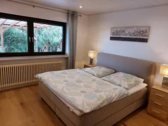 Ein gemütliches Schlafzimmer mit einem großen Bett und zwei Nachttischen. Durch das Fenster fällt natürlicher Lichtschein auf den Holzfußboden.