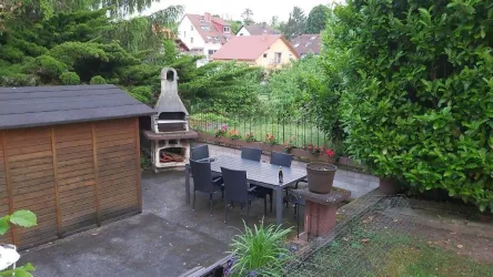 Ein schöner Garten mit einer Terrasse und einem Esstisch mit Stühlen. Im Hintergrund sind ein Grill und viel grüne Vegetation zu sehen.