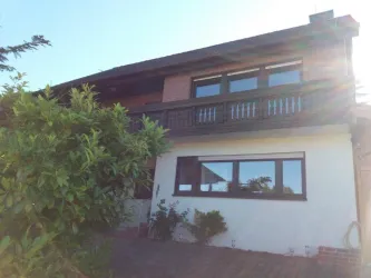 Ein modernes Haus mit einem Balkon und großen Fenstern. Es ist von Pflanzen und einem Garten umgeben.