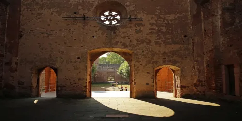Ein historischer Innenraum mit mehreren Bögen und einer rauen, steinernen Wand. Durch die Öffnungen sind grüne Bäume und Licht zu sehen.